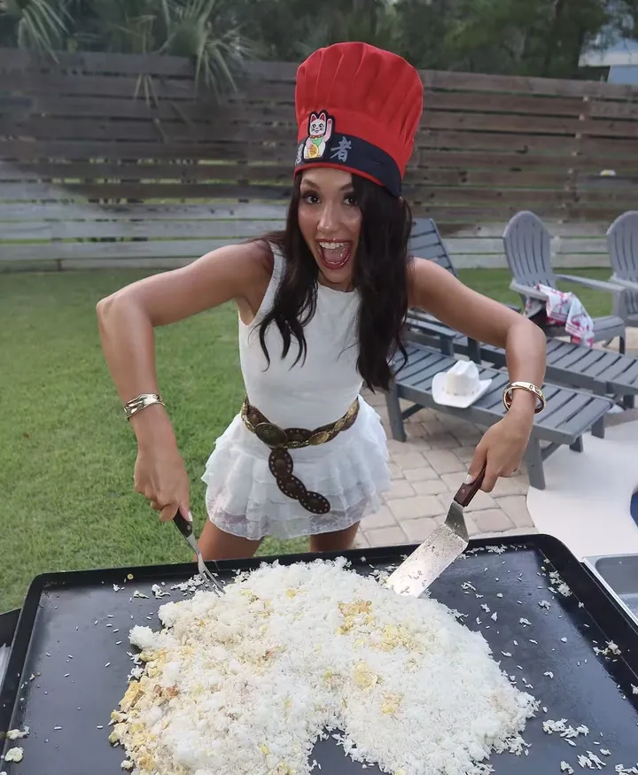 Guest in a red chef hat holding spatulas over fried rice on a hibachi grill