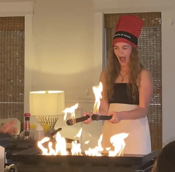 Guest wearing a red chef hat holding cooking tools above flames at the grill